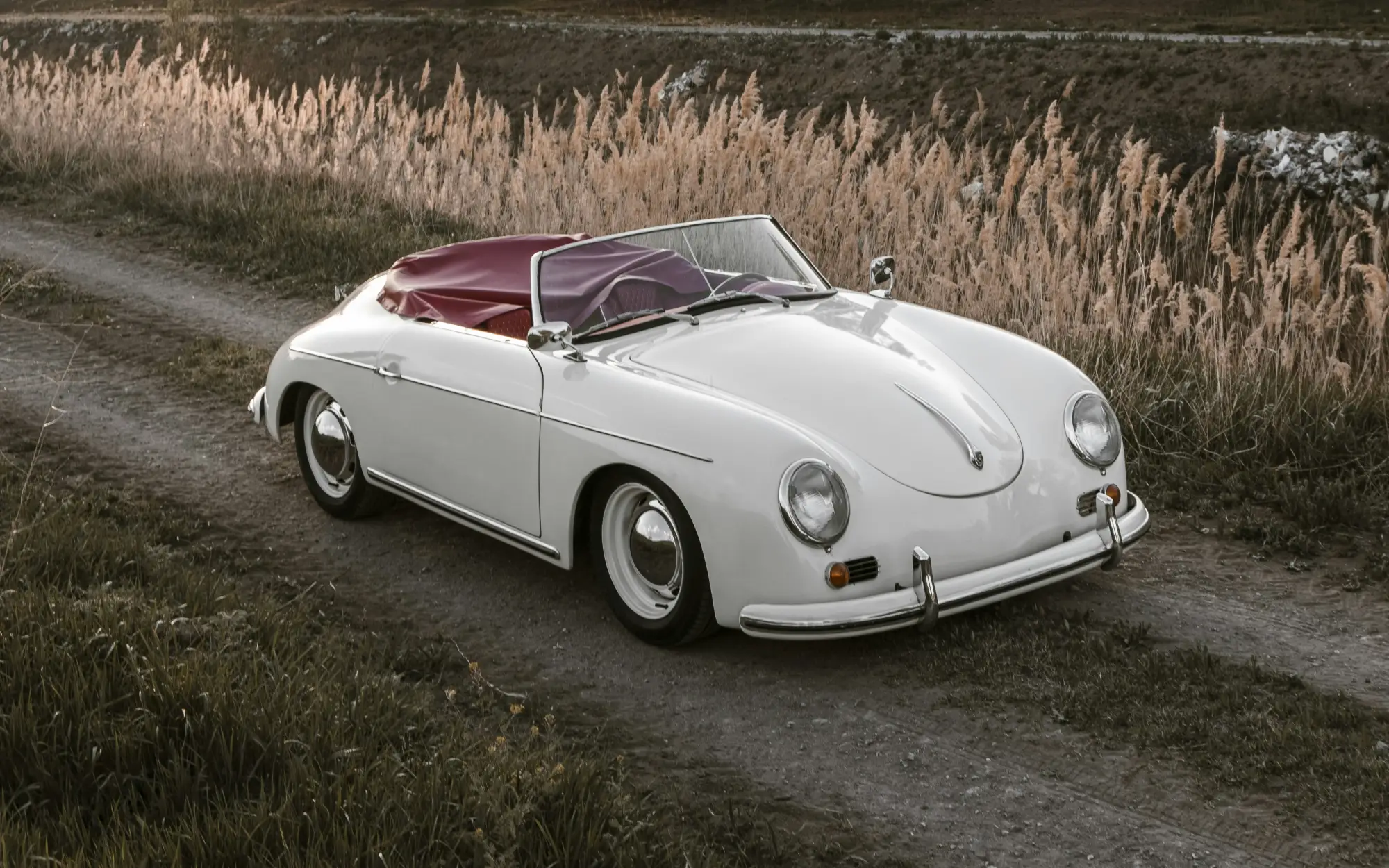 White Porsche 356 convertible with a red top on a country road beside tall reeds